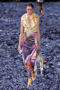 Model walks a fashion show wearing a floral, ruffled top and bright, printed pants with fabric scarves wrapped at the waist, on a blue textured runway.