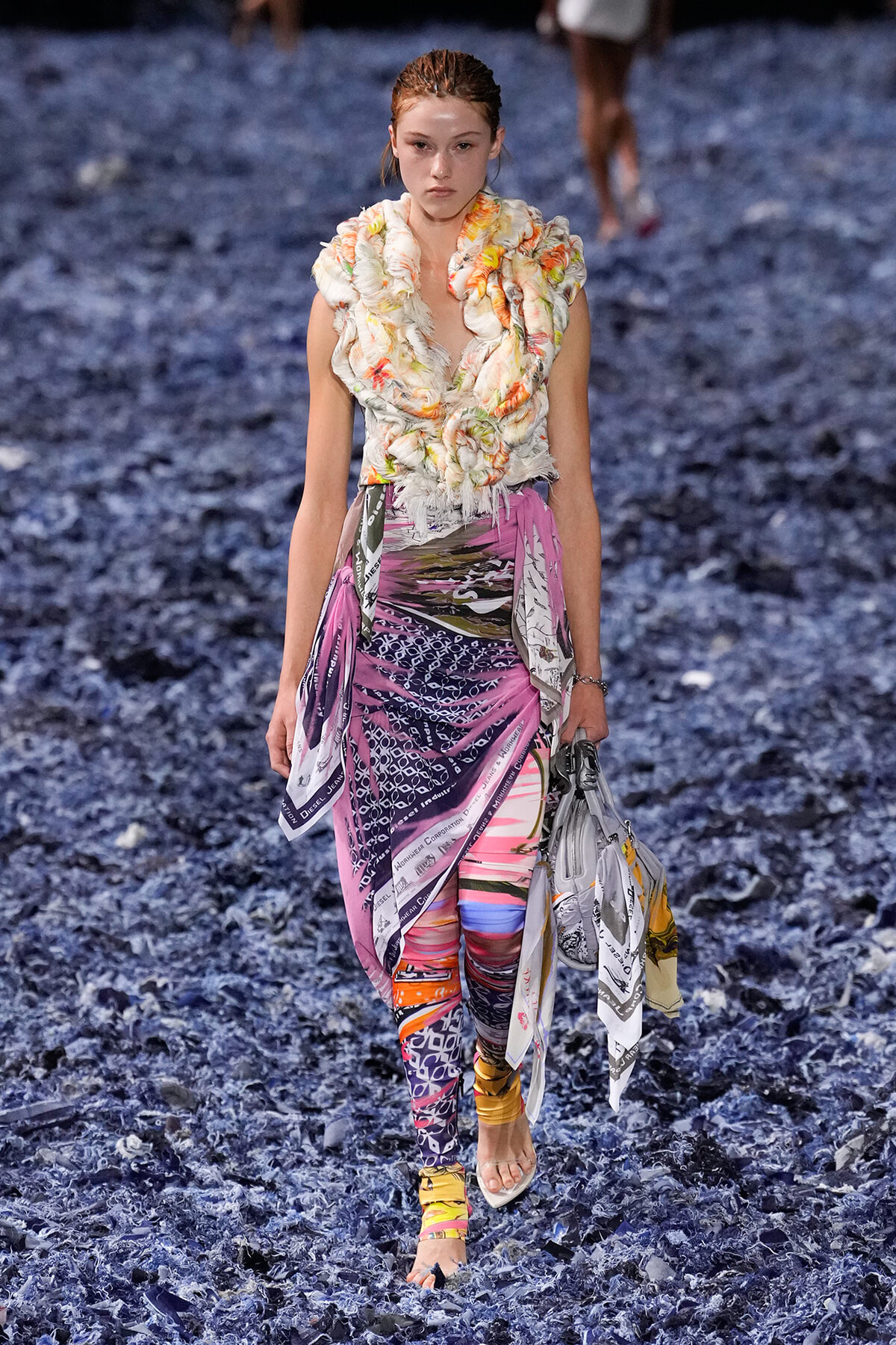Model walks a fashion show wearing a floral, ruffled top and bright, printed pants with fabric scarves wrapped at the waist, on a blue textured runway.