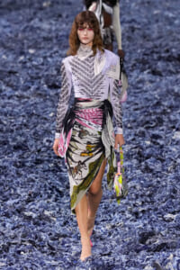 Model walking a runway in a lavender geometric-pattern top and a wrapped, multicolored print skirt on a blue shredded-floor set.