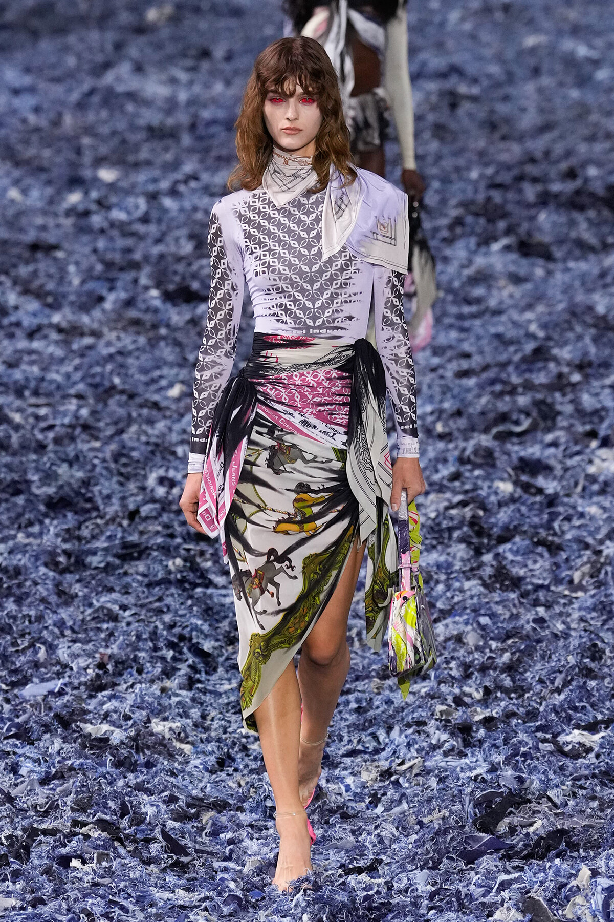 Model walking a runway in a lavender geometric-pattern top and a wrapped, multicolored print skirt on a blue shredded-floor set.