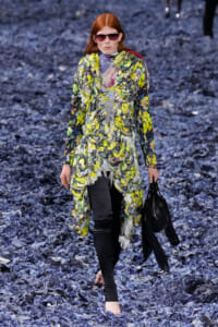 Model walking a runway in a vibrant yellow-green floral coat over a patterned top, with red sunglasses and a black bag.