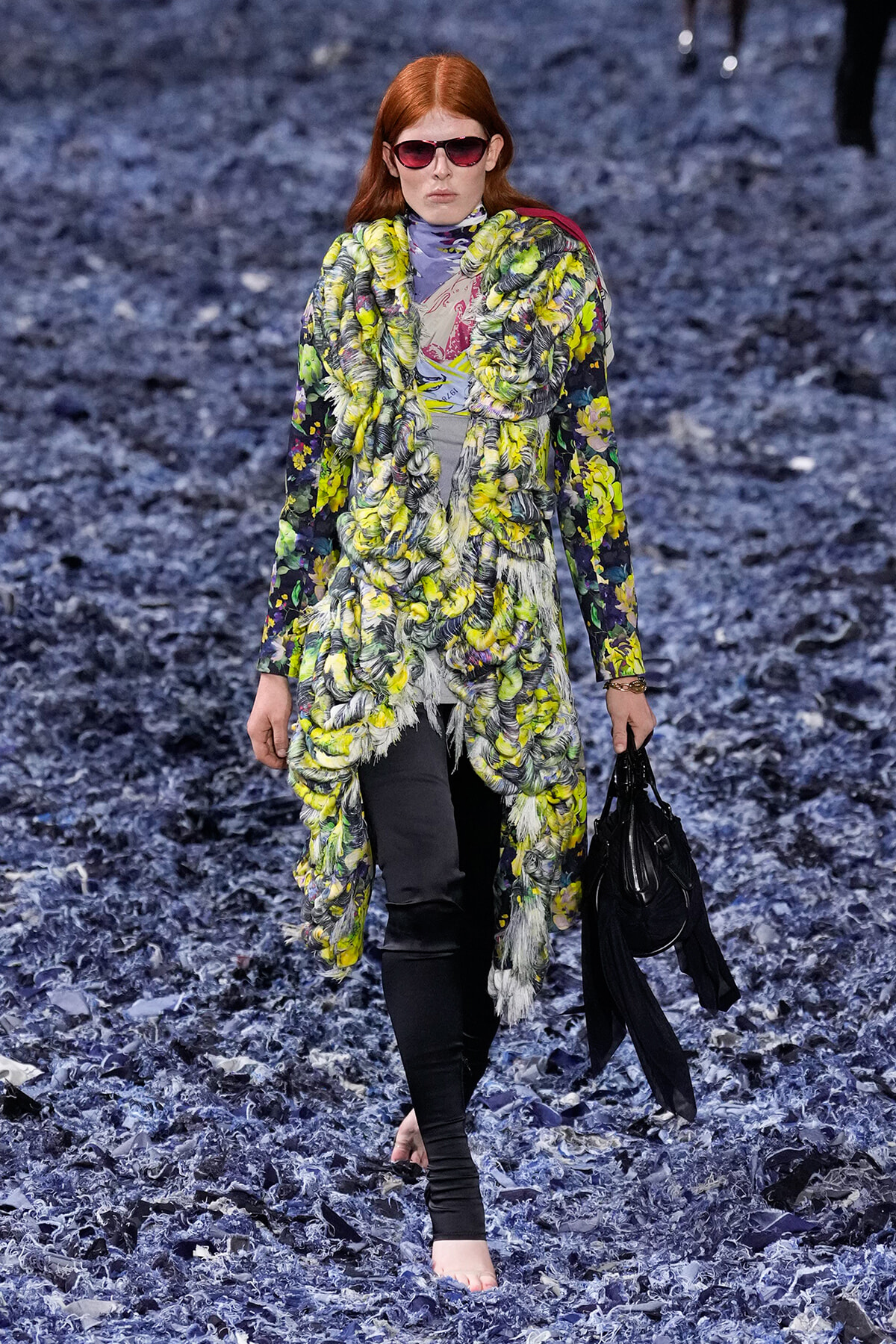 Model walking a runway in a vibrant yellow-green floral coat over a patterned top, with red sunglasses and a black bag.