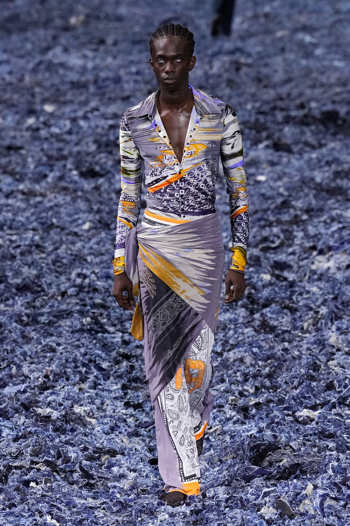 Male model walks a fashion show wearing a purple-gray patterned shirt and a matching wrap skirt with orange accents on a blue-toned, leaf-covered runway