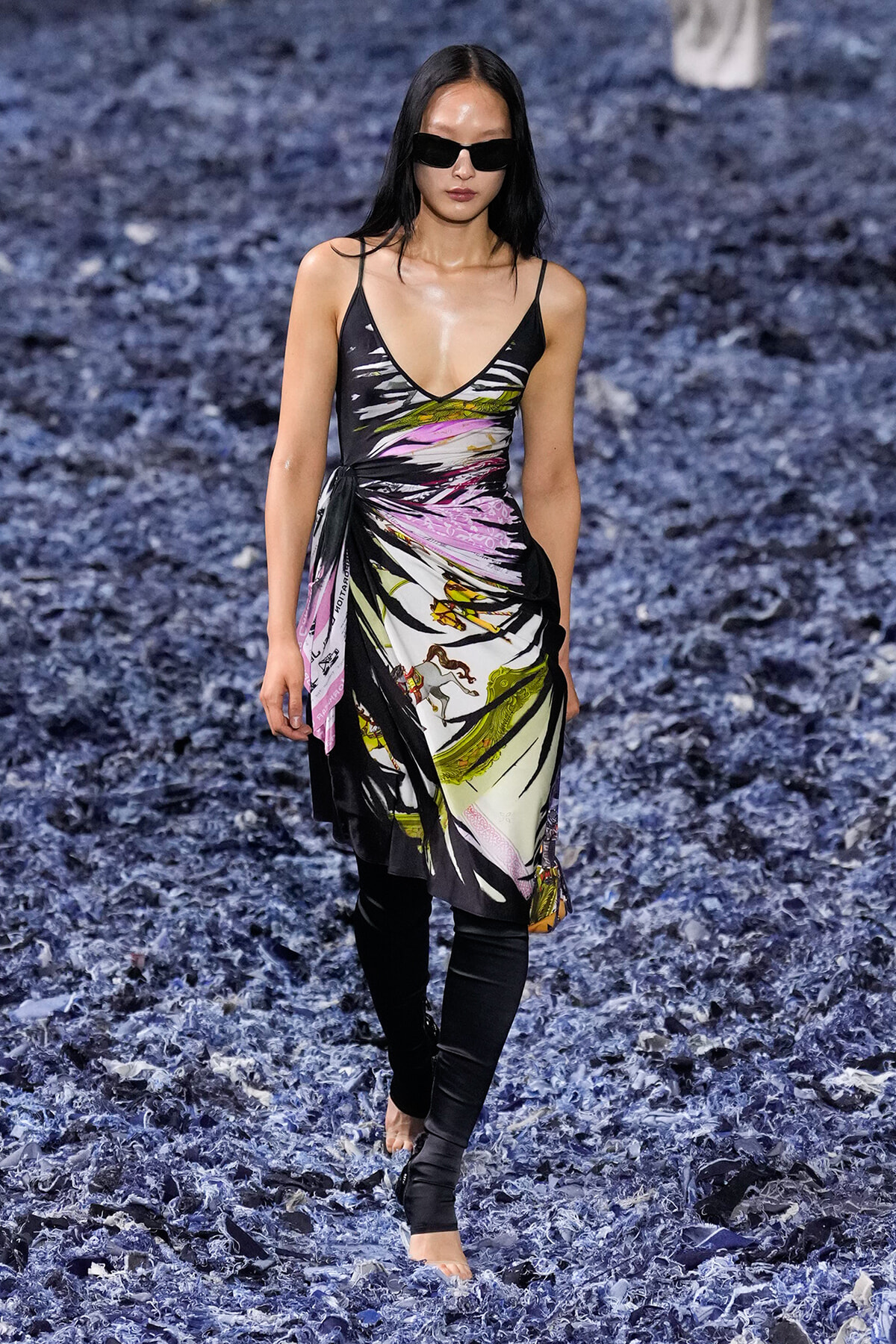 Model walks the runway in a colorful, abstract-printed slip dress with black leggings and dark sunglasses.