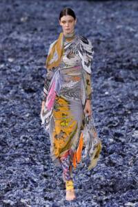 Model walks a fashion runway in a long, multicolored printed wrap dress with scarf details, barefoot on a blue textured runway surface.