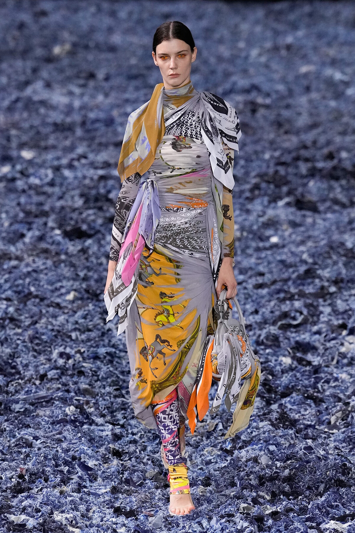 Model walks a fashion runway in a long, multicolored printed wrap dress with scarf details, barefoot on a blue textured runway surface.
