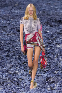 Model walking a runway in a gray printed mini dress with red sash, carrying patterned bags against a blue textured runway backdrop.