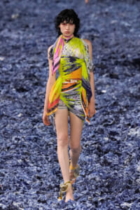Model walking on a blue rocky surface wearing a neon multicolor wrap dress with a scarf-like tie at the neck and matching sandals.