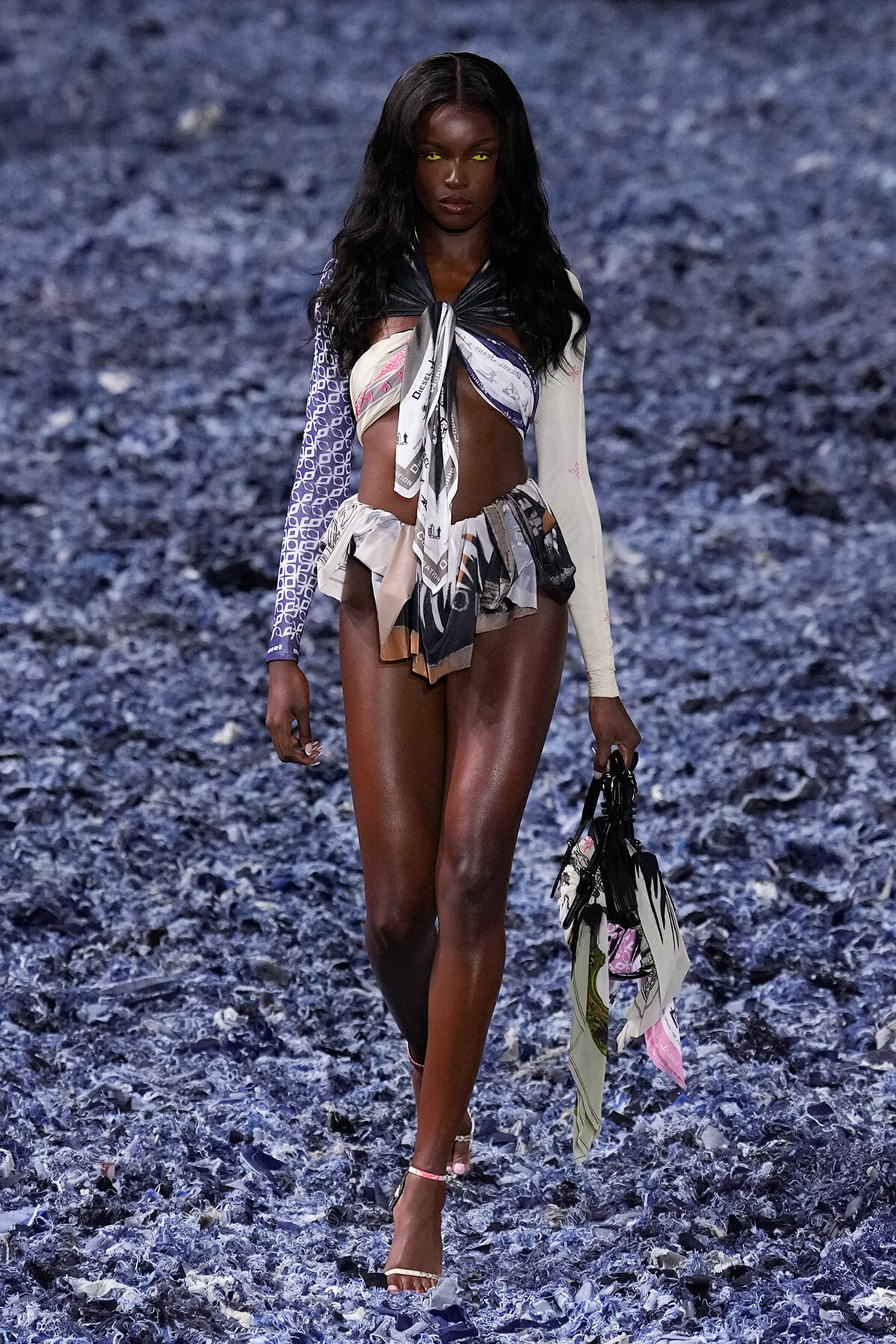 Model walking a fashion runway on a blue textured floor, wearing a knot-front scarf top with one purple patterned sleeve and printed wrap shorts, holding a scarfed bag.