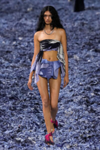 Model walks a runway in a strapless black-and-white bandeau with a tied scarf detail and blue layered shorts, on a blue confetti runway with pink sandals.