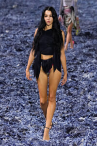 Model walking a runway on a blue textured floor, wearing a black wrap top and bikini bottoms with straps.