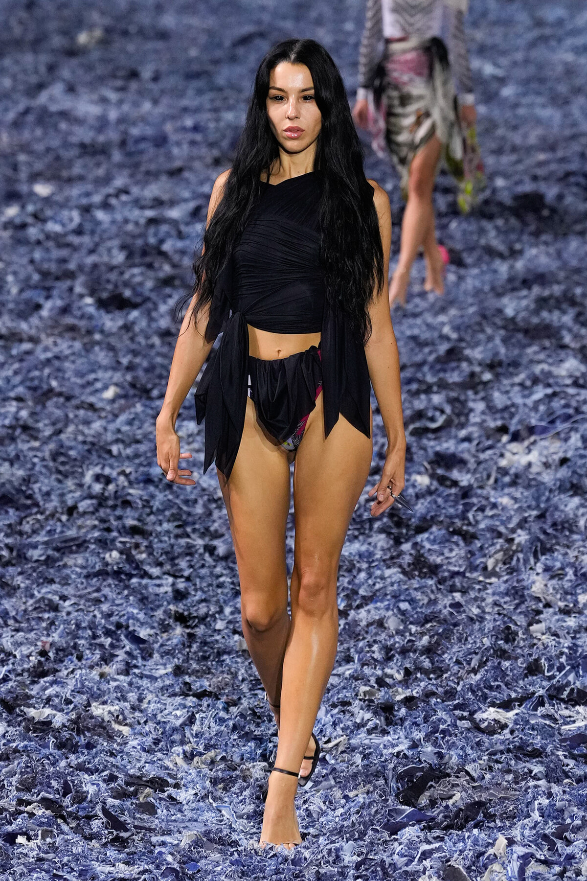 Model walking a runway on a blue textured floor, wearing a black wrap top and bikini bottoms with straps.