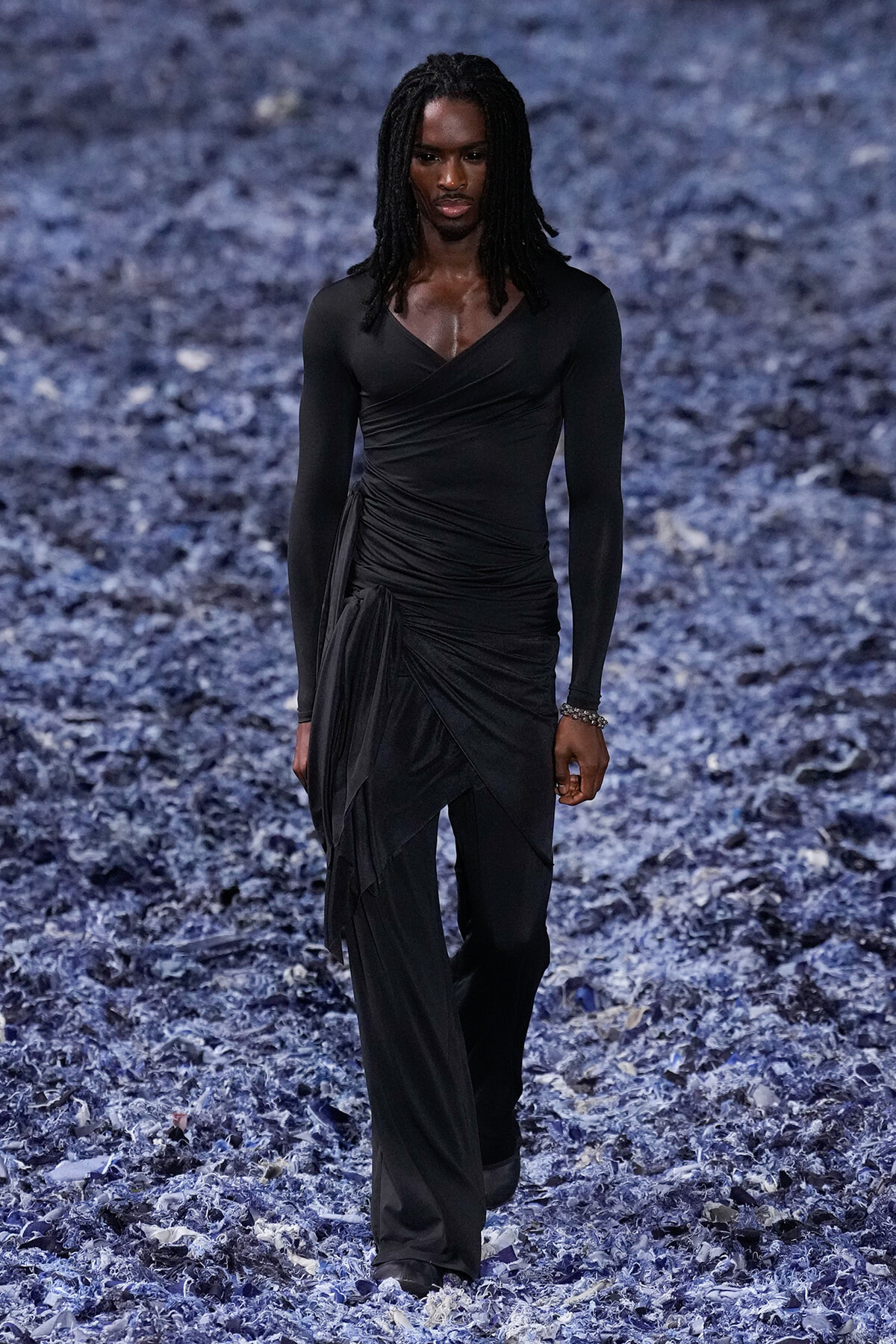 Full-body shot of a model wearing a black draped, wrap-style outfit with long sleeves, walking on a blue-toned runway surface.
