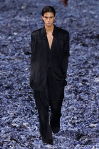 Male model wearing a loose black suit with an open shirt walks down a blue-gray textured runway.