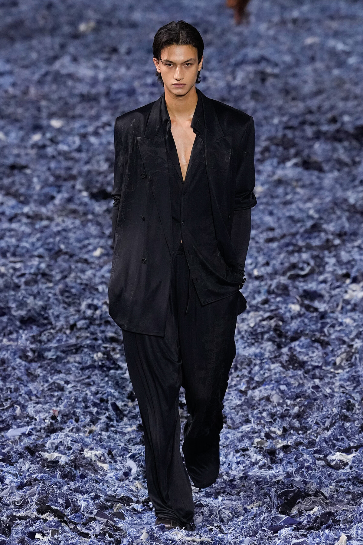 Male model wearing a loose black suit with an open shirt walks down a blue-gray textured runway.
