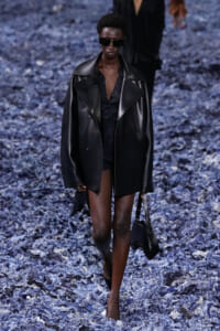 Model walking a fashion runway in a black leather oversized coat, black top, shorts, and sunglasses on a blue-petal runway backdrop.