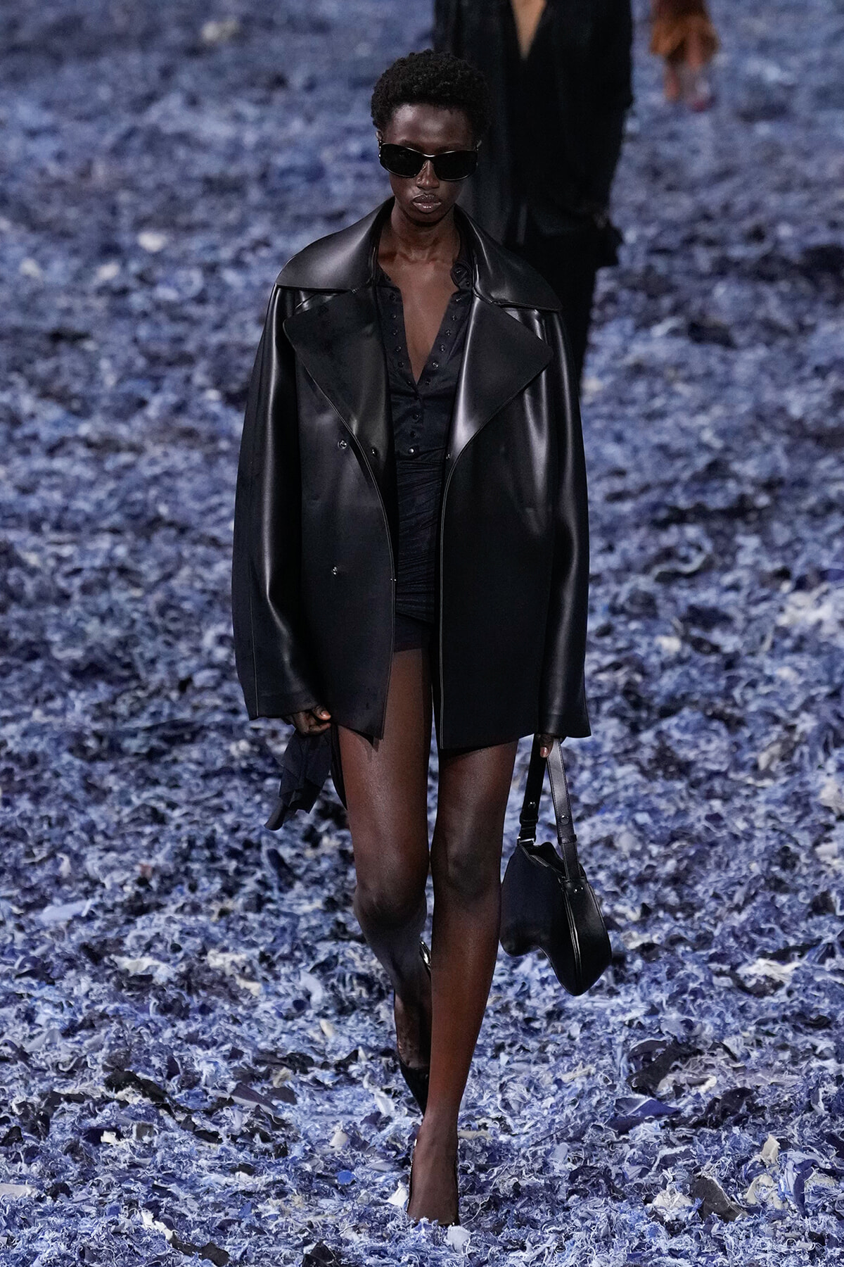 Model walking a fashion runway in a black leather oversized coat, black top, shorts, and sunglasses on a blue-petal runway backdrop.