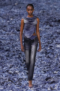 Model walks the runway in a metallic blue draped top and matching gray pants on a blue textured backdrop.