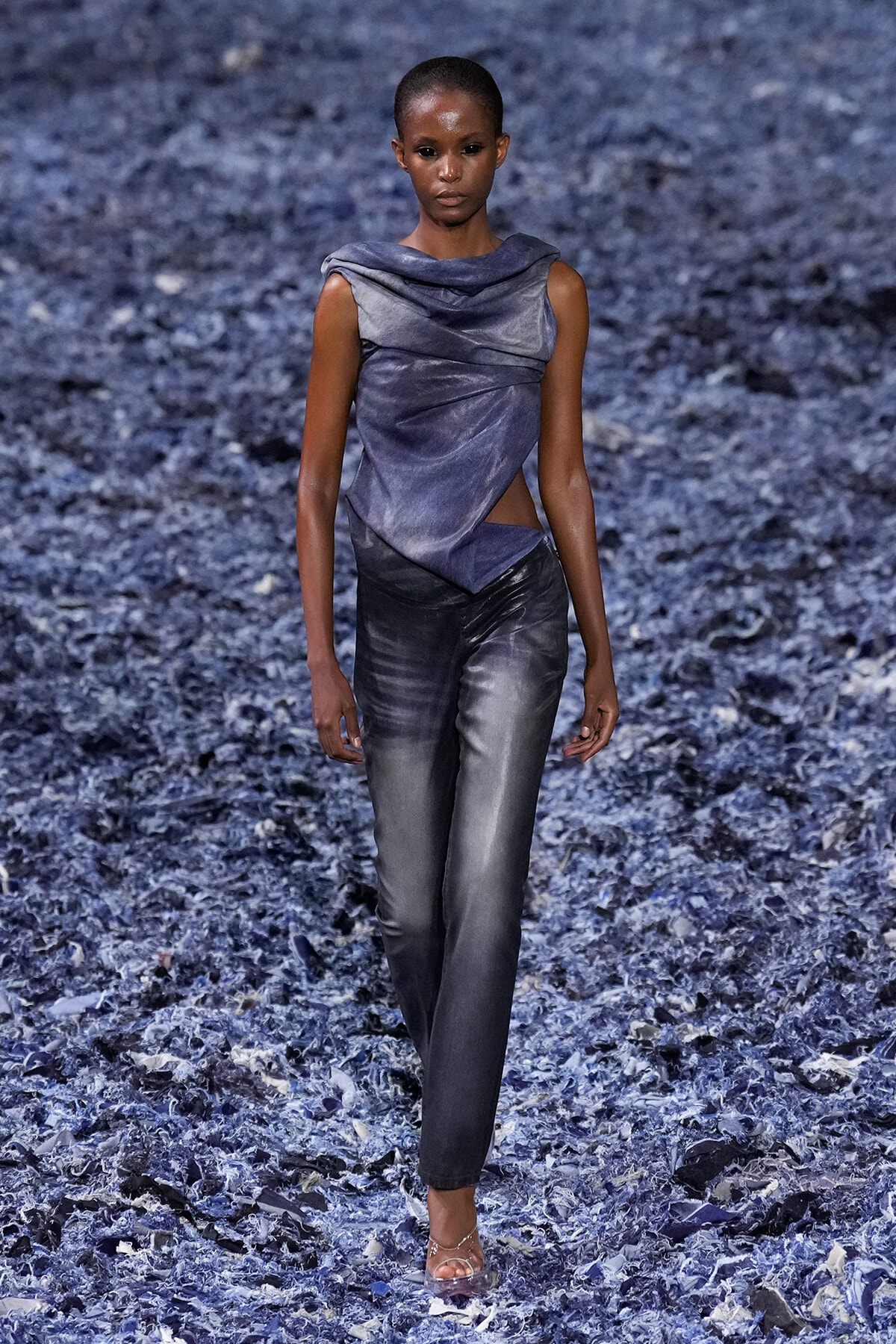 Model walks the runway in a metallic blue draped top and matching gray pants on a blue textured backdrop.