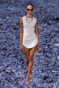 Female model walking a runway in a sleeveless white mini dress with layered silver necklaces and dark sunglasses.