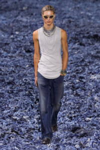 Male model walking on a blue-toned runway wearing a sleeveless white top with layered necklaces and sunglasses, paired with dark jeans and bracelets.