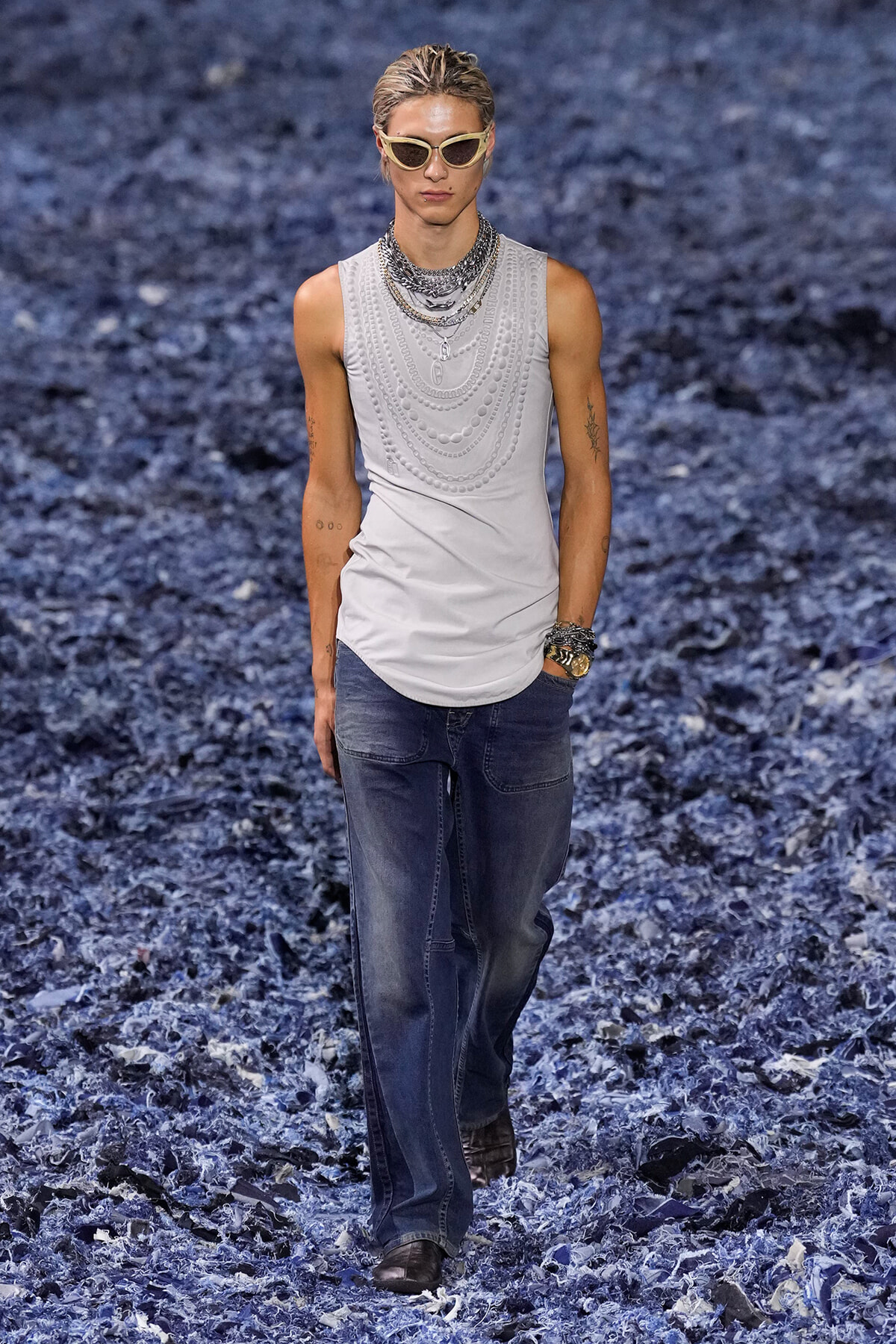 Male model walking on a blue-toned runway wearing a sleeveless white top with layered necklaces and sunglasses, paired with dark jeans and bracelets.