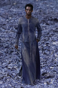 Male model wearing a long blue denim robe-dress walking on a rocky blue-gray landscape.