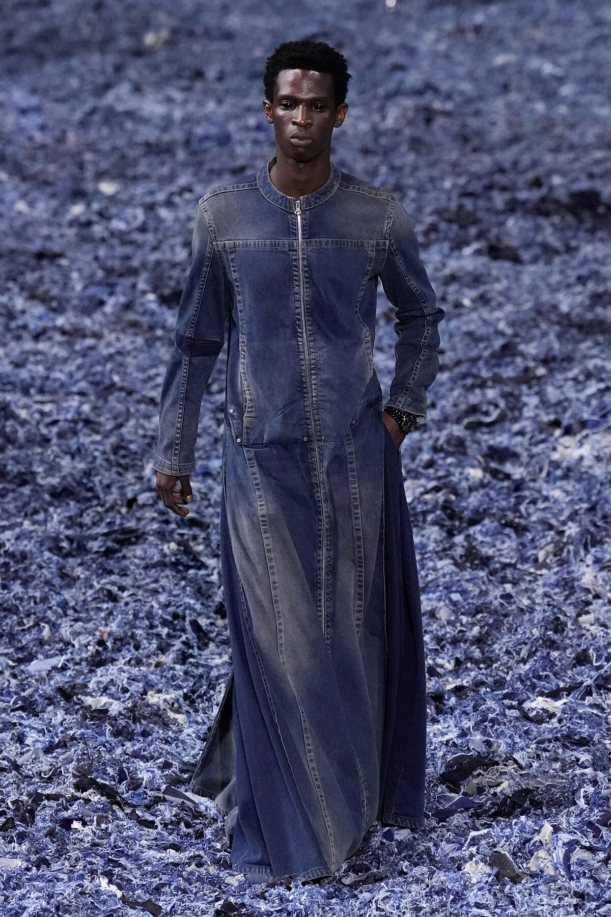 Male model wearing a long blue denim robe-dress walking on a rocky blue-gray landscape.