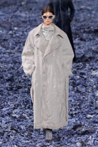 Model walking the runway in a oversized distressed beige trench coat with white sunglasses and scarf, on a blue-textured catwalk surface.