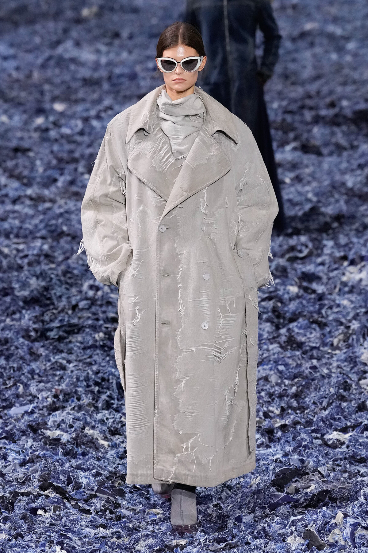 Model walking the runway in a oversized distressed beige trench coat with white sunglasses and scarf, on a blue-textured catwalk surface.