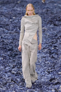 Model walking a runway in a light gray distressed jumpsuit with cutouts, standing on a dark blue textured floor. Yellow contact lenses create a striking gaze.