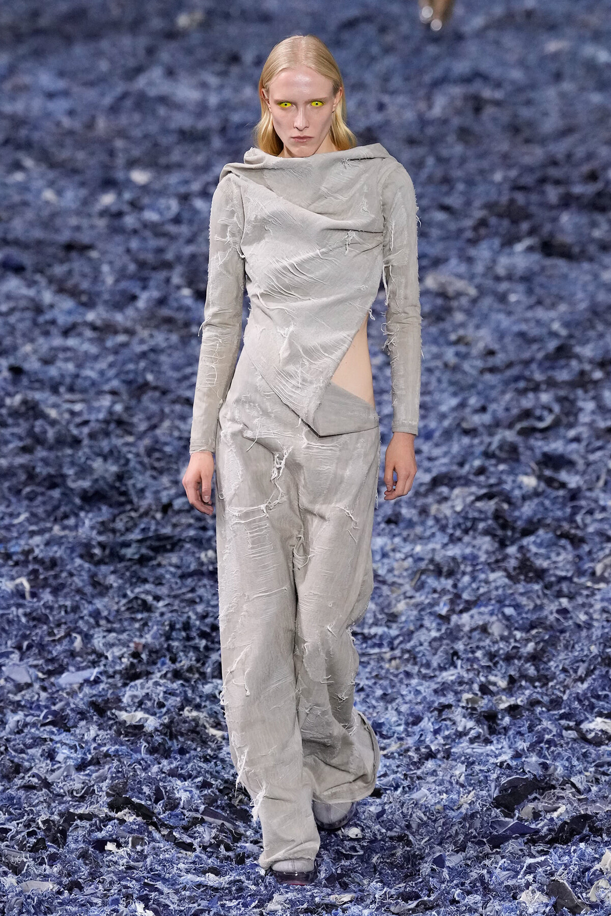 Model walking a runway in a light gray distressed jumpsuit with cutouts, standing on a dark blue textured floor. Yellow contact lenses create a striking gaze.