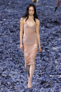 Model walking down a runway in a pale pink, form-fitting dress with cut-out panels; blue shredded floor decor.
