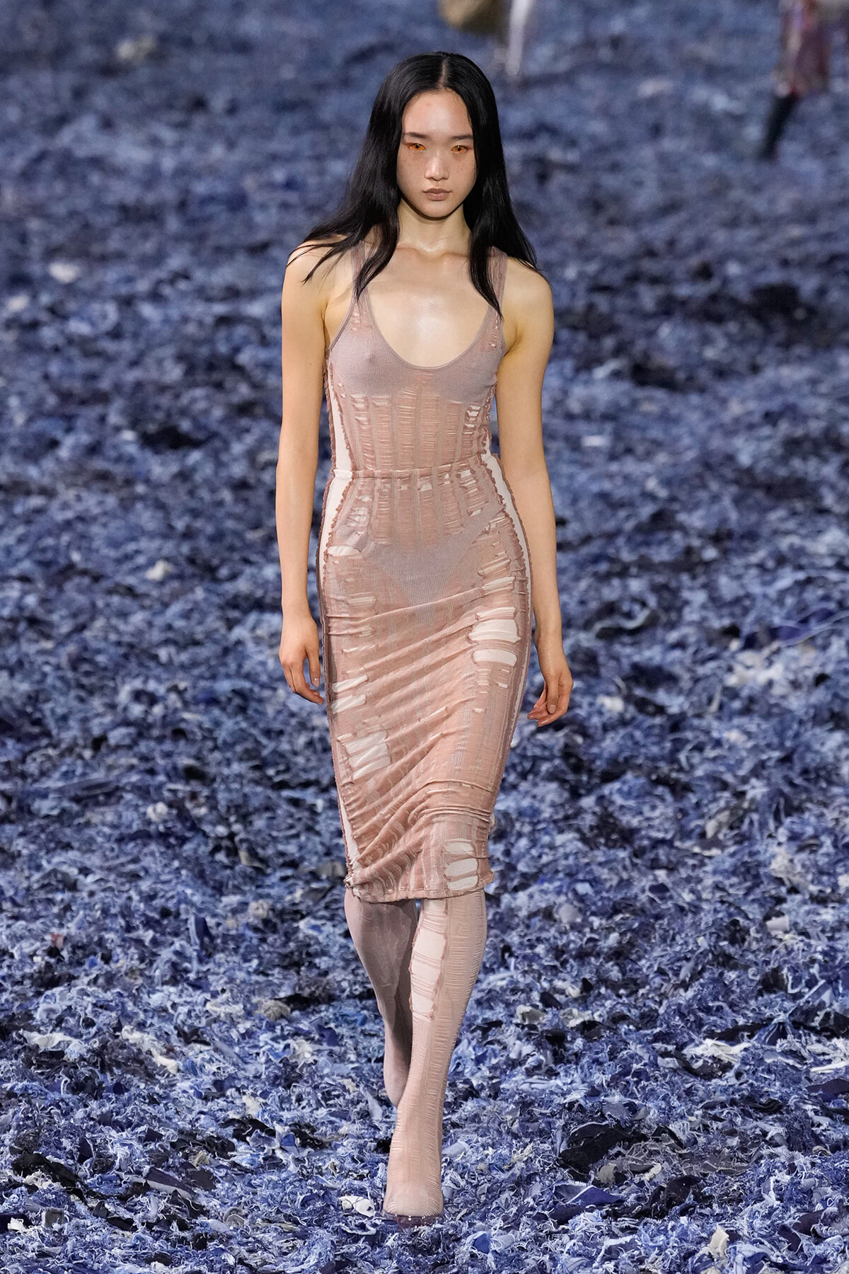 Model walking down a runway in a pale pink, form-fitting dress with cut-out panels; blue shredded floor decor.