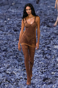 Model walks down a runway in a brown sheer bodysuit with striped panels, blue-flecked floor beneath her.