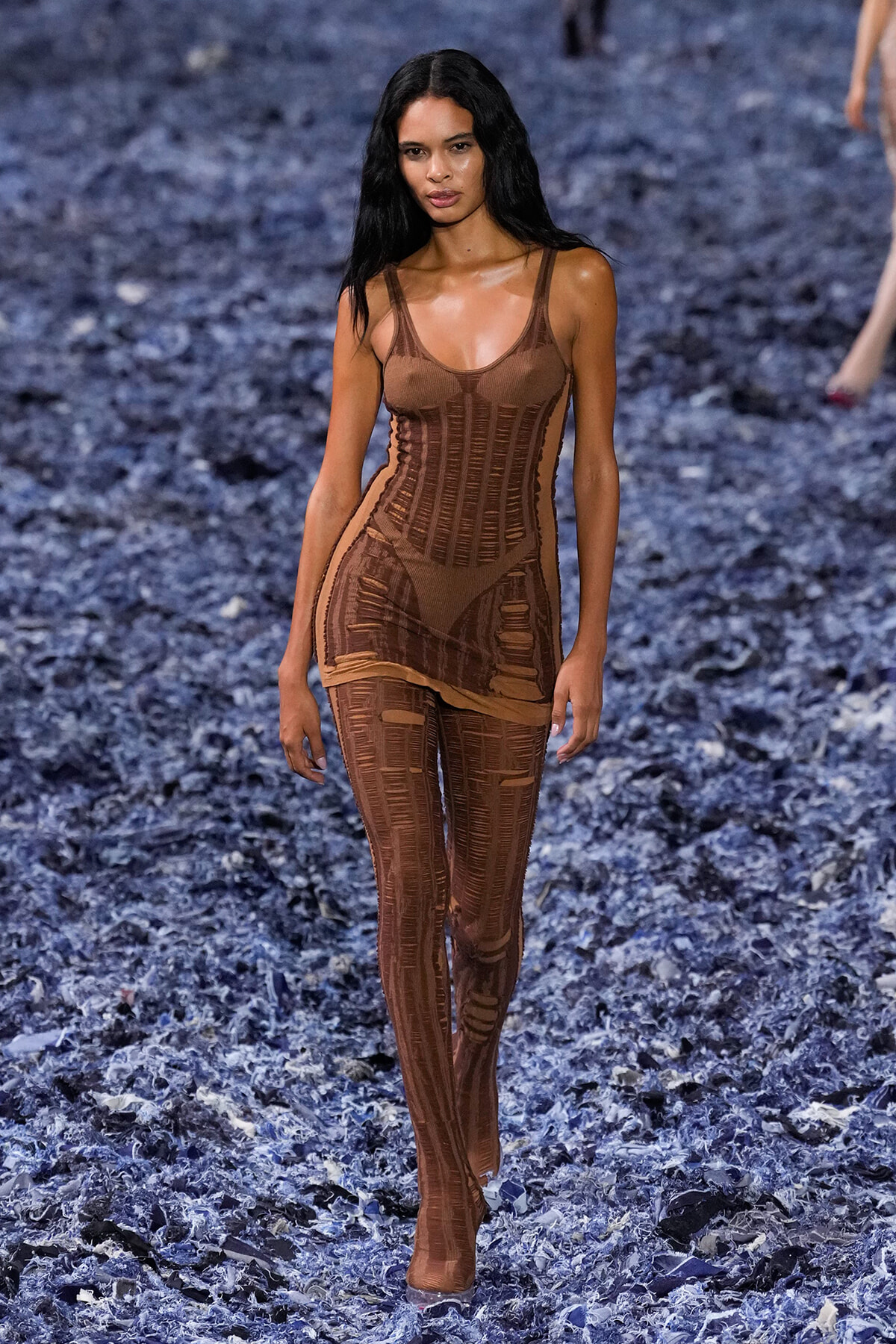 Model walks down a runway in a brown sheer bodysuit with striped panels, blue-flecked floor beneath her.