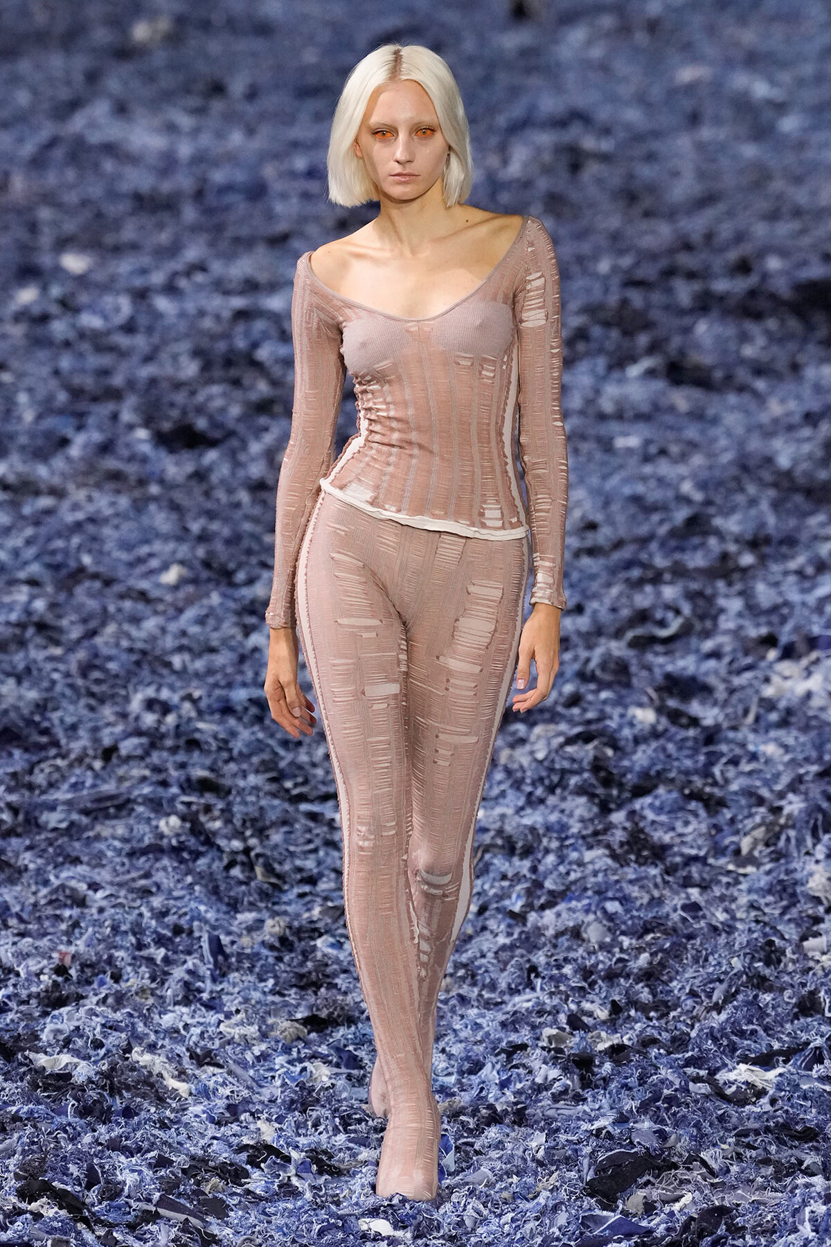 Model walking a runway in a pale pink sheer, distressed-knit bodysuit and leggings set.