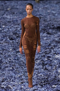 Model walking a runway in a brown, sheer, form‑fitting dress with vertical panels and cutouts against a blue textured floor.