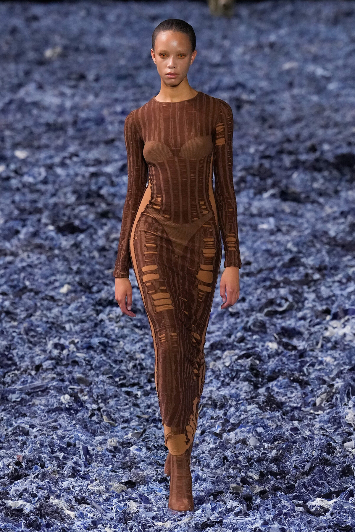 Model walking a runway in a brown, sheer, form‑fitting dress with vertical panels and cutouts against a blue textured floor.