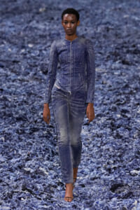 Female model walking a runway in a bluewashed denim outfit with a zip-front jacket and matching jeans against a rocky blue backdrop.