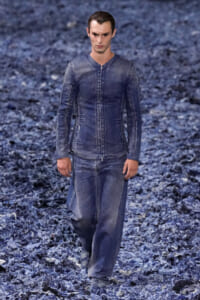 Male model walking a runway in a blue distressed denim jacket and matching wide-leg jeans, with a blue textured backdrop.