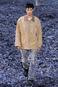 Male model walks the runway in a beige distressed zip jacket and ripped jeans against a blue textured floor background.