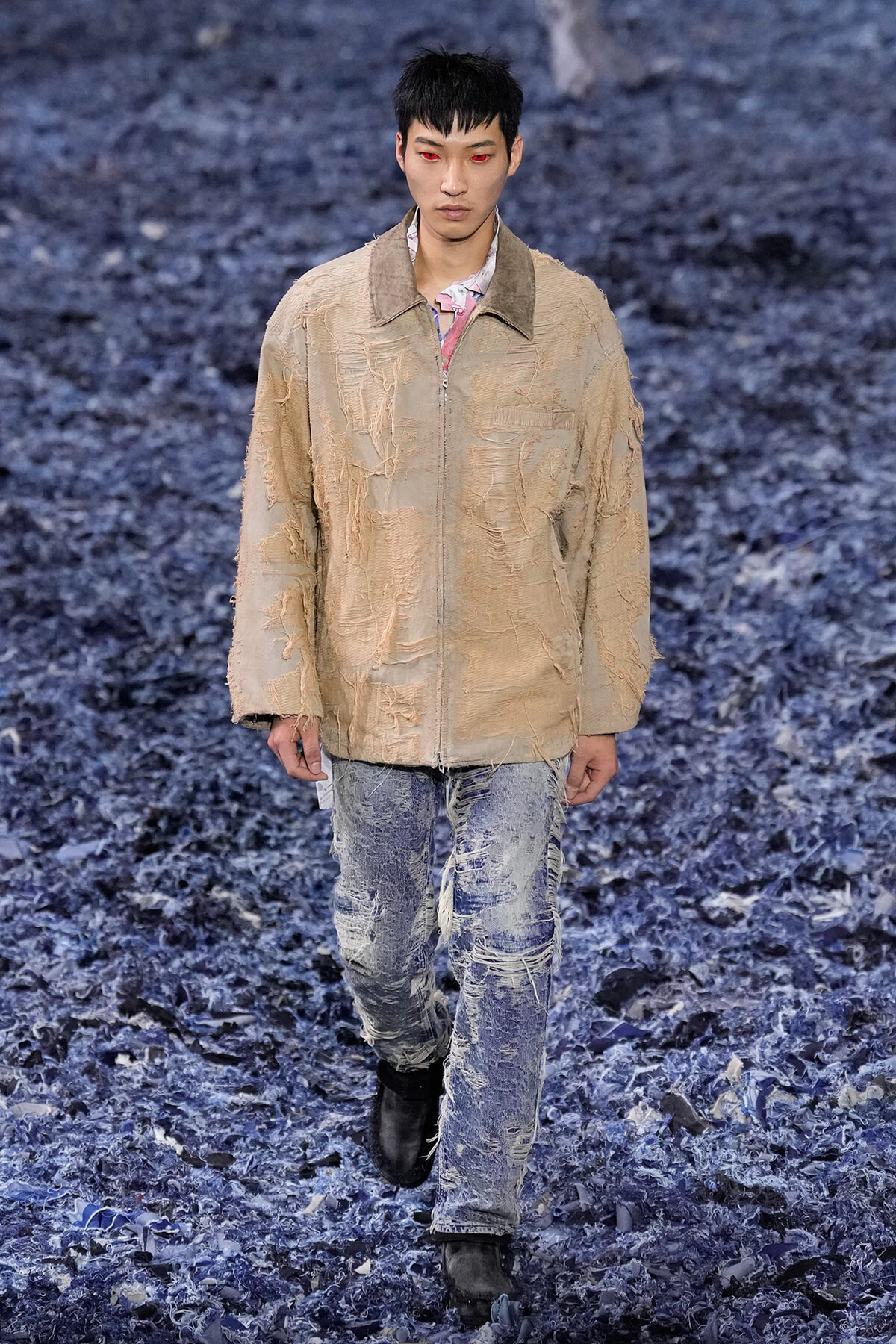 Male model walks the runway in a beige distressed zip jacket and ripped jeans against a blue textured floor background.