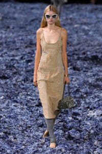 Model walking a runway in a distressed beige slip dress, white cat-eye sunglasses, gray leg warmers, and a gray handbag on a blue-textured runway.