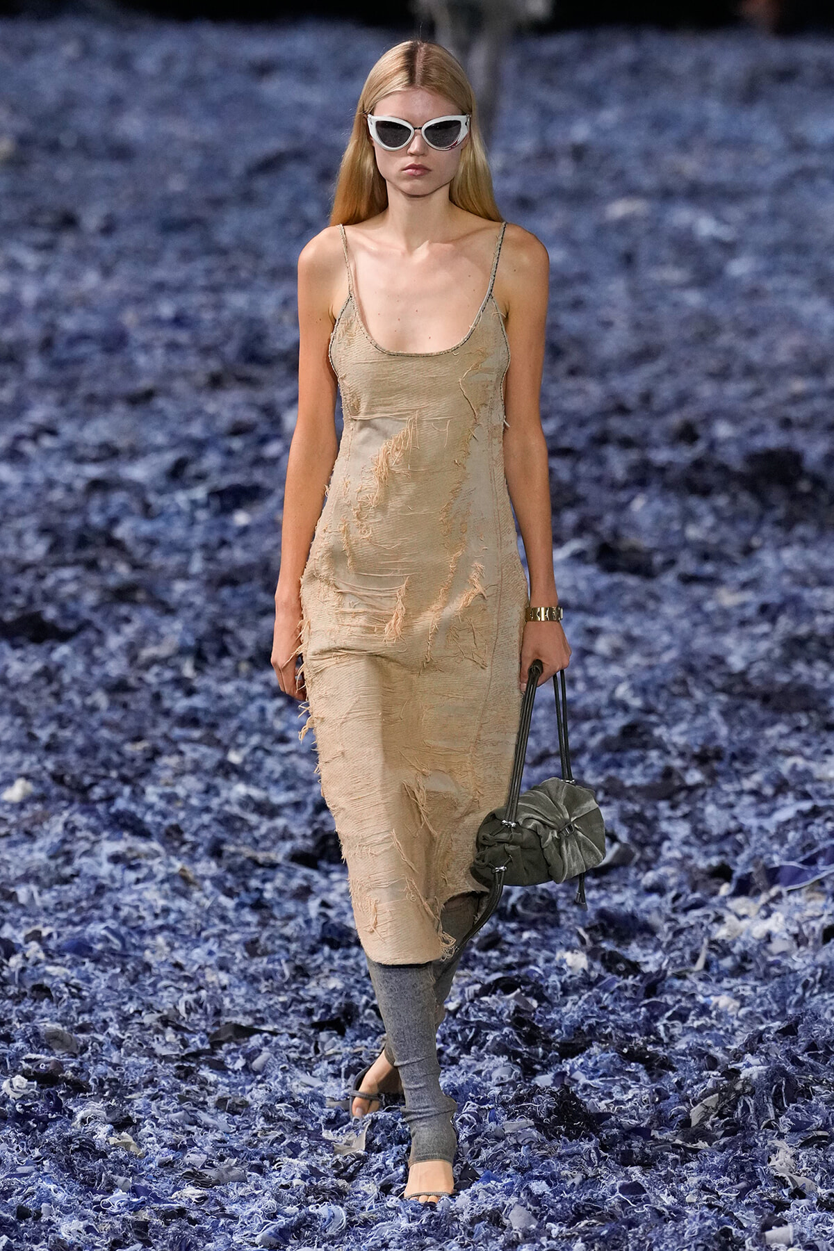 Model walking a runway in a distressed beige slip dress, white cat-eye sunglasses, gray leg warmers, and a gray handbag on a blue-textured runway.