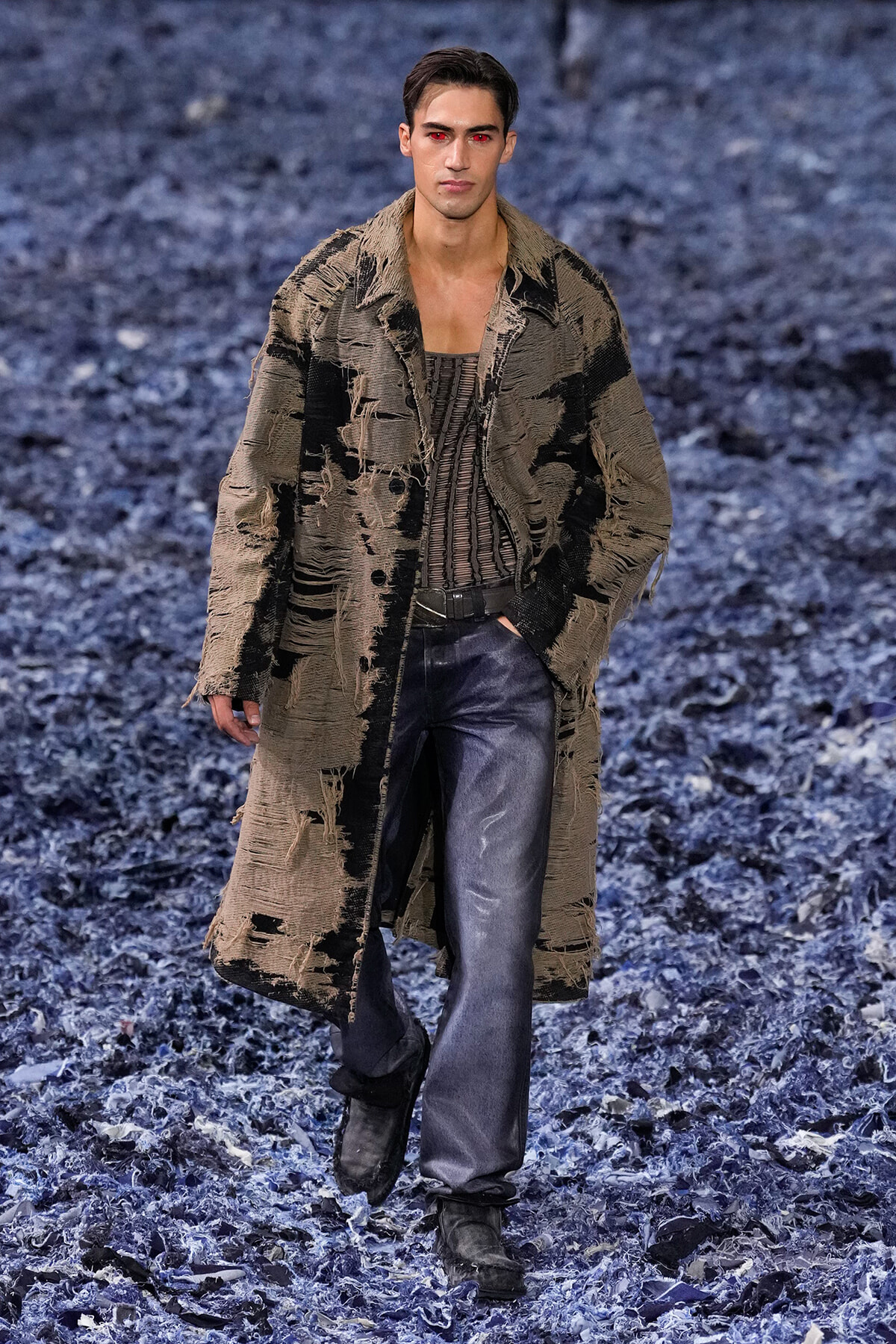 Male model walking a fashion show wearing a distressed beige-brown long coat over a striped top and dark jeans on a blue-toned runway.