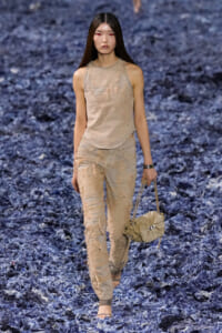 Model walking a fashion runway in distressed beige sleeveless top and matching ripped pants, barefoot, carrying a beige handbag.