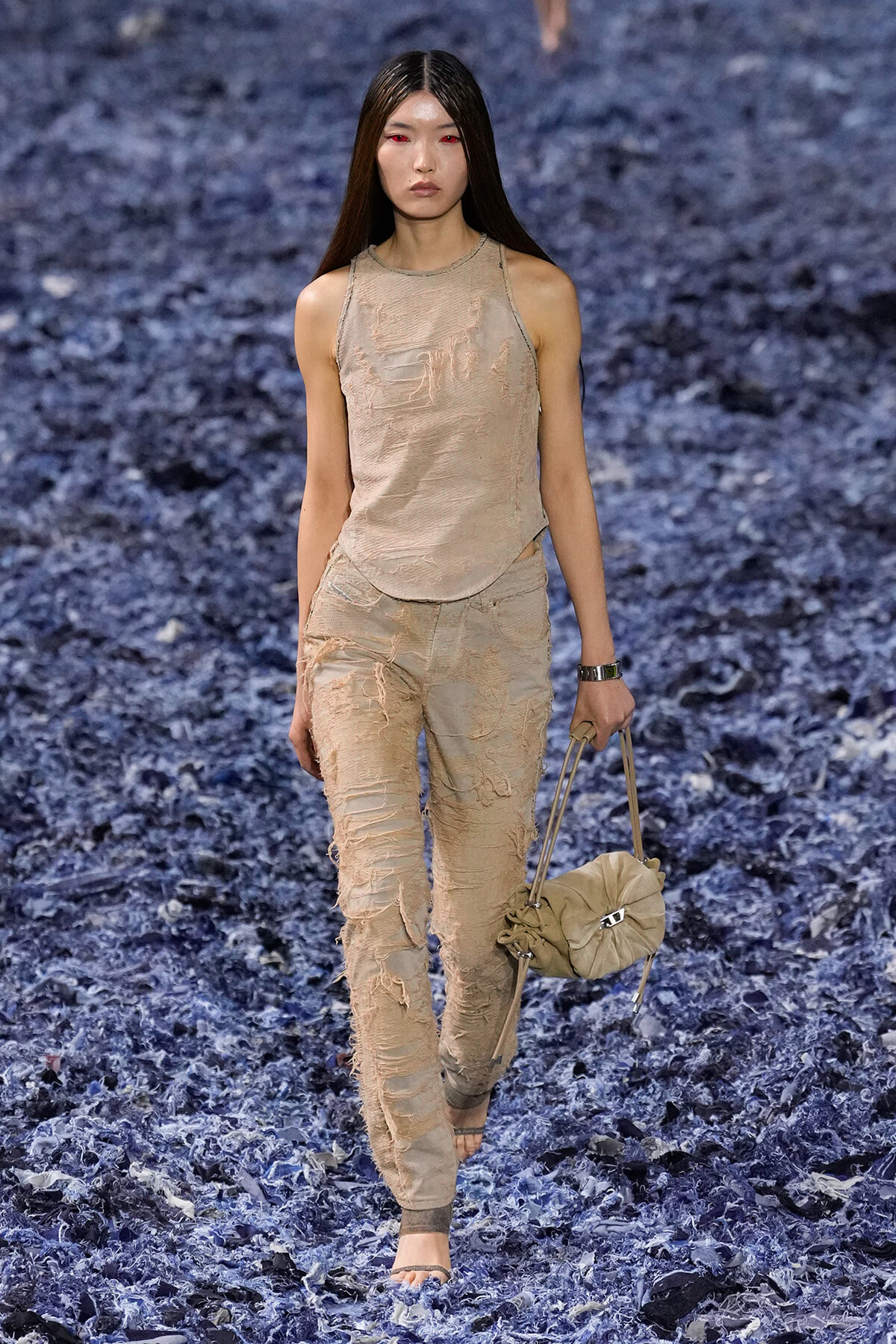 Model walking a fashion runway in distressed beige sleeveless top and matching ripped pants, barefoot, carrying a beige handbag.