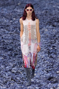 Model walking a runway in a sheer white sleeveless top with graphic text, colorful patterned pants, and gray knee-high boots wearing white sunglasses.