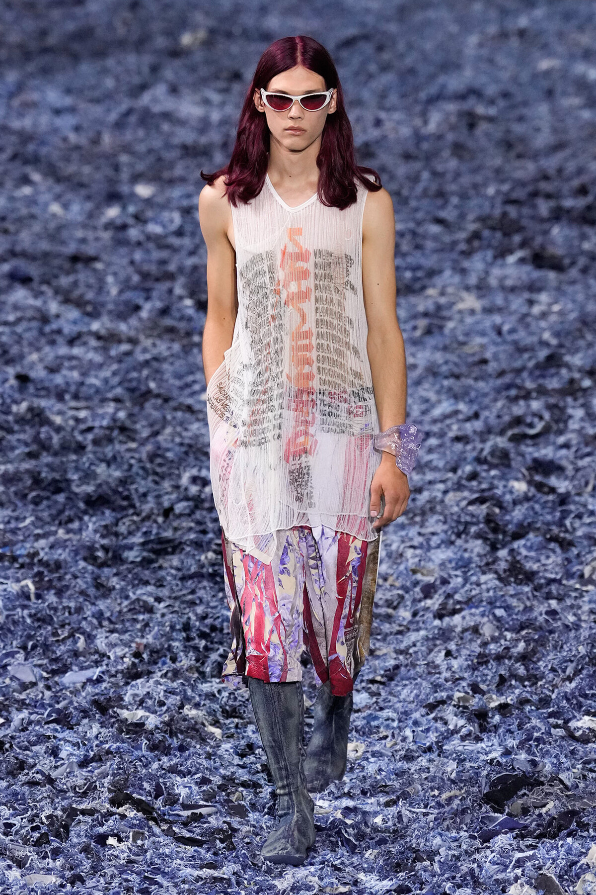 Model walking a runway in a sheer white sleeveless top with graphic text, colorful patterned pants, and gray knee-high boots wearing white sunglasses.